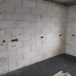 Wnętrze w stanie surowym z widoczną instalacją elektryczną: białe przewody w peszlach i pomarańczowe puszki pod gniazdka, zamontowane na ścianach z betonu komórkowego.