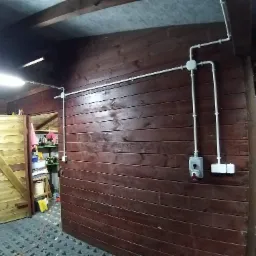 Wewnętrzna instalacja elektryczna natynkowa w garażu lub warsztacie z drewnianymi ścianami, widoczne białe rury i puszki instalacyjne oraz wyłączniki.