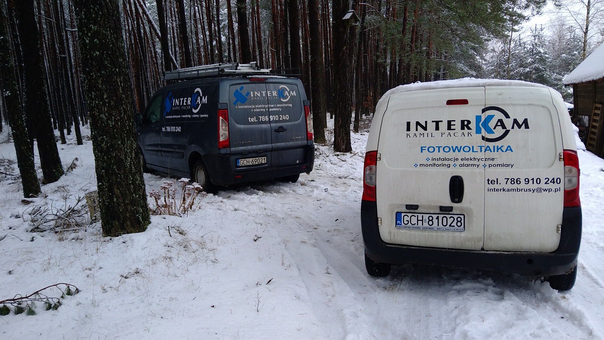 Dwa firmowe samochody dostawcze z logo firmy Interkam Kamil Pacak zaparkowane na ośnieżonej drodze w lesie. Na białym samochodzie napis Fotowoltaika oraz instalacje elektryczne, monitoring, alarmy...