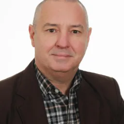 Portret mężczyzny w brązowej marynarce i koszuli w kratę na białym tle. Uśmiechnięty, z kr&oacute;tkimi włosami i widocznymi zmarszczkami wok&oacute;ł oczu.