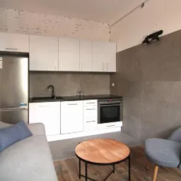 Nowoczesna kuchnia z białymi szafkami, czarnym blatem i sprzętem AGD ze stali nierdzewnej, połączona z częścią dzienną z szarą sofą i drewnianym stolikiem kawowym.