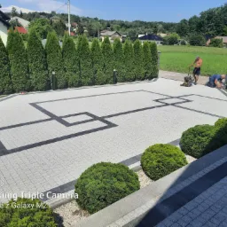 Dwóch mężczyzn przy pracy na świeżo ułożonym podjeździe z kostki brukowej w geometryczny wzór, jeden zagęszcza powierzchnię mechanicznym ubijakiem, drugi koryguje krawędzie.