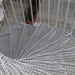 Spiralne schody zewnętrzne z metalowej siatki, widok z góry, otoczone jasną nawierzchnią i roślinami w donicach.