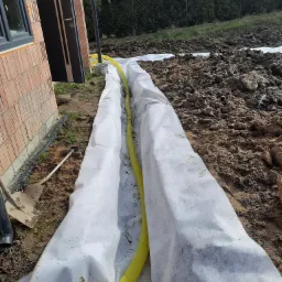 Fundament budynku w trakcie izolacji: żółta rura drenażowa ułożona w wykopie wyłożonym białą geowłókniną, widoczna ceglana ściana budynku i narzędzia.