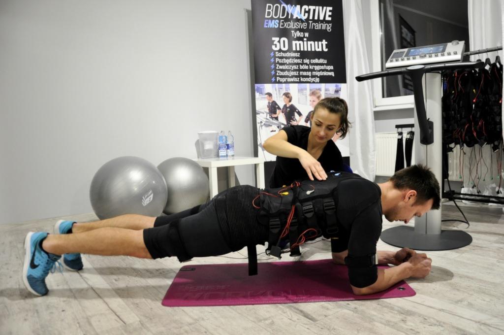 Mężczyzna w pozycji plank z elektrostymulacją EMS, asystowany przez kobietę, w tle piłki fitness i plakat informacyjny o treningu EMS.