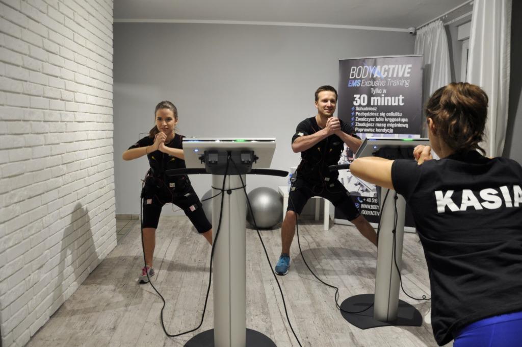 Dwoje ćwiczących w kombinezonach EMS przed urządzeniami do elektrostymulacji mięśni, trenerka z napisem KASIA na plecach obserwuje trening, w tle ściana z cegły i plakat reklamowy Body Active.