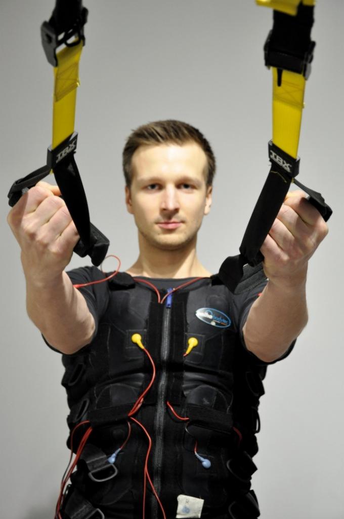 Mężczyzna w czarnym kombinezonie EMS z podpiętymi kablami, trzymający w dłoniach żółte taśmy TRX, prezentuje ćwiczenie podczas treningu w Gdyni.