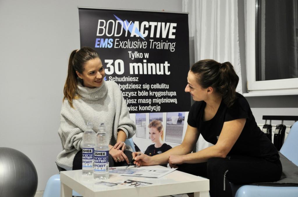 Dwie kobiety omawiają plan treningowy EMS, na stole butelki wody mineralnej, w tle baner reklamowy BODYACTIVE EMS Exclusive Training.
