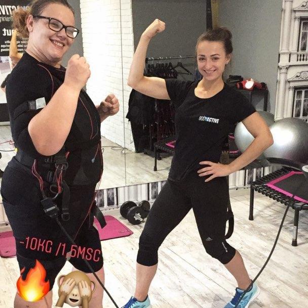 Dwie kobiety w strojach do elektrostymulacji mięśni (EMS) w siłowni w Gdyni, prezentują efekty treningu, z widocznym napisem '-10KG / 1,5MSC'.