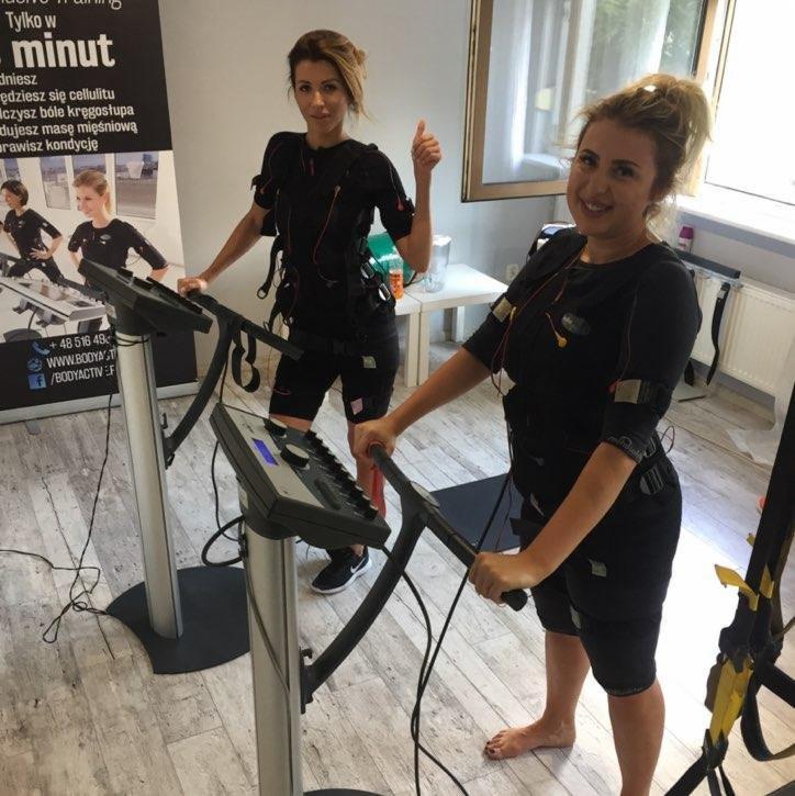 Dwie kobiety uśmiechają się podczas treningu EMS na bieżniach; jedna z nich pokazuje kciuk w górę, a druga stoi boso.