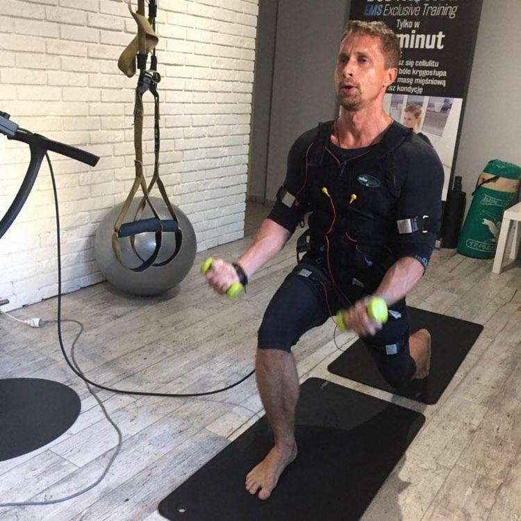 Mężczyzna w kombinezonie EMS wykonuje wykroki z małymi hantlami w dłoniach, na czarnej macie do ćwiczeń, w tle ściana z białej cegły i plakat reklamowy EMS.
