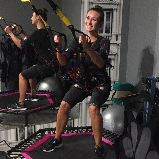 Kobieta uśmiecha się podczas ćwiczeń EMS na trampolinie z gumami oporowymi w studio fitness. W tle druga osoba wykonuje to samo ćwiczenie.