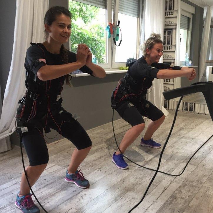 Dwie kobiety w strojach do elektrostymulacji mięśni wykonują ćwiczenia w jasnym pomieszczeniu z parkietem i oknem w tle.