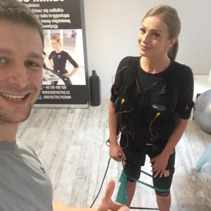 Uśmiechnięta kobieta w kombinezonie EMS z elektrodami, pozująca w studio, obok uśmiechnięty mężczyzna robiący selfie z uniesionym kciukiem, w tle plakat reklamowy z informacjami kontaktowymi.