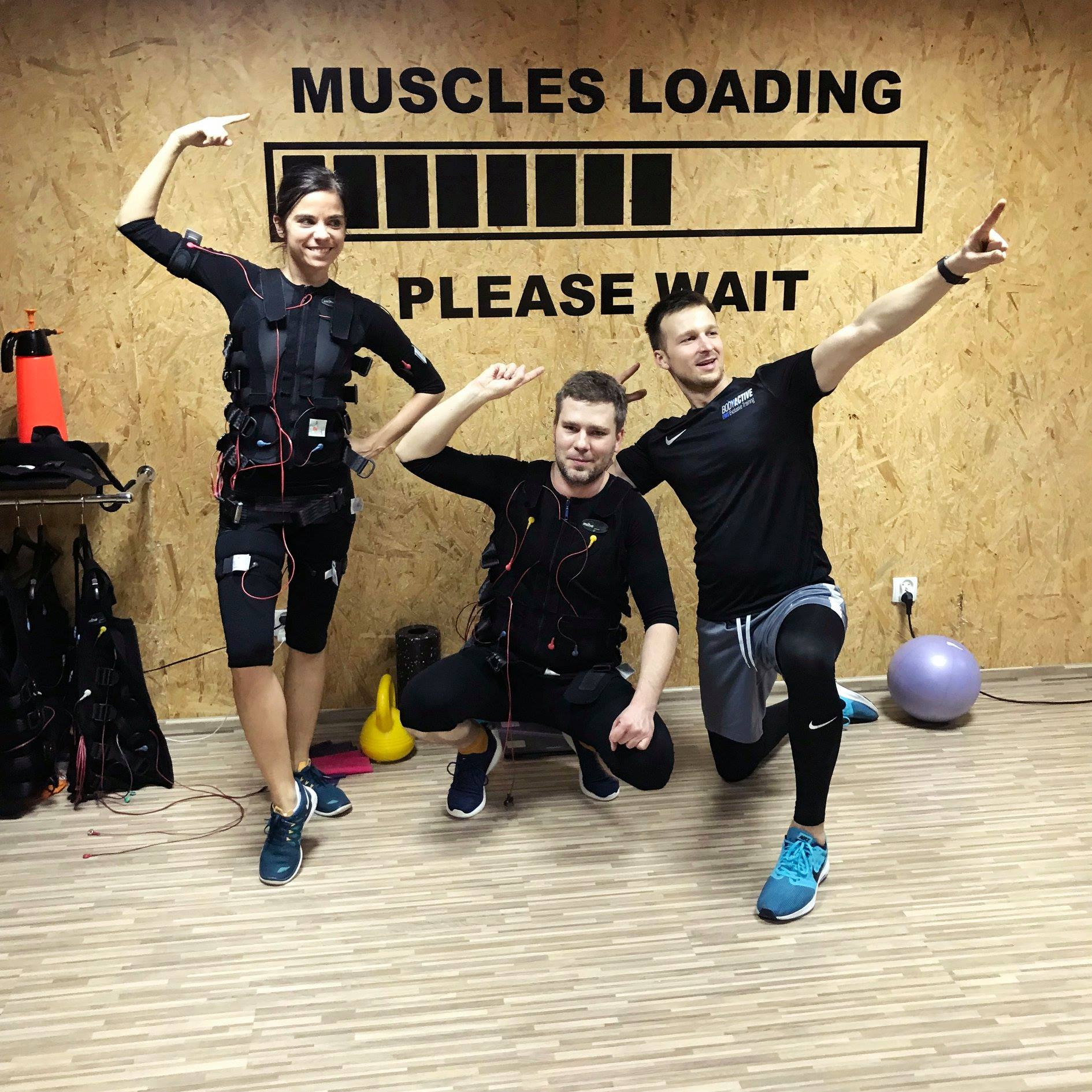 Trzy osoby w strojach do elektrostymulacji mięśni pozują w studiu fitness z napisem 'MUSCLES LOADING' na drewnianej ścianie.