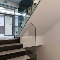 Szklana balustrada zamocowana na metalowych uchwytach, zabezpieczająca schody z ciemnego kamienia w nowoczesnym wnętrzu.