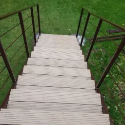 Zewnętrzne schody z drewnopodobnych desek kompozytowych, z brązowymi metalowymi poręczami z poziomymi prętami, widok z góry w kierunku zielonego trawnika.