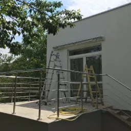 Montaż balustrady ze stali nierdzewnej na balkonie, widoczne dwie drabiny, narzędzia i zwinięty wąż na podłodze, w tle biała ściana budynku i zieleń drzew.