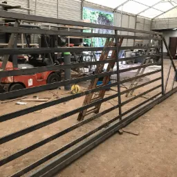 Surowa, metalowa brama wjazdowa w trakcie produkcji w warsztacie, widoczny wózek widłowy i drabina w tle.