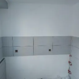 Ściana w trakcie układania płytek ceramicznych w jasnych odcieniach szarości, widoczne krzyżyki dystansowe i puszki elektryczne.