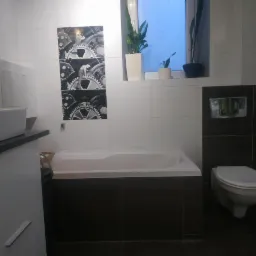 Nowoczesna łazienka z białą ceramiką, czarnymi akcentami i dekoracyjnymi płytkami z motywem kół zębatych. Rośliny w doniczkach na parapecie okna dodają naturalnego elementu.