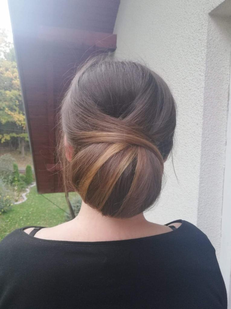 Upięty kok z brązowych włosów z pasemkami w odcieniach blondu, widok z tyłu, na tle elewacji budynku i zieleni.