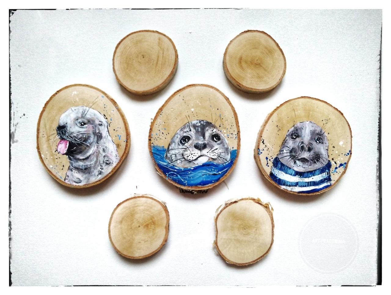 Ręcznie malowane, artystyczne plasterki drewna z wizerunkami fok: jedna z wyciągniętym językiem, druga w wodzie, trzecia w marynarskim swetrze; na białym tle.