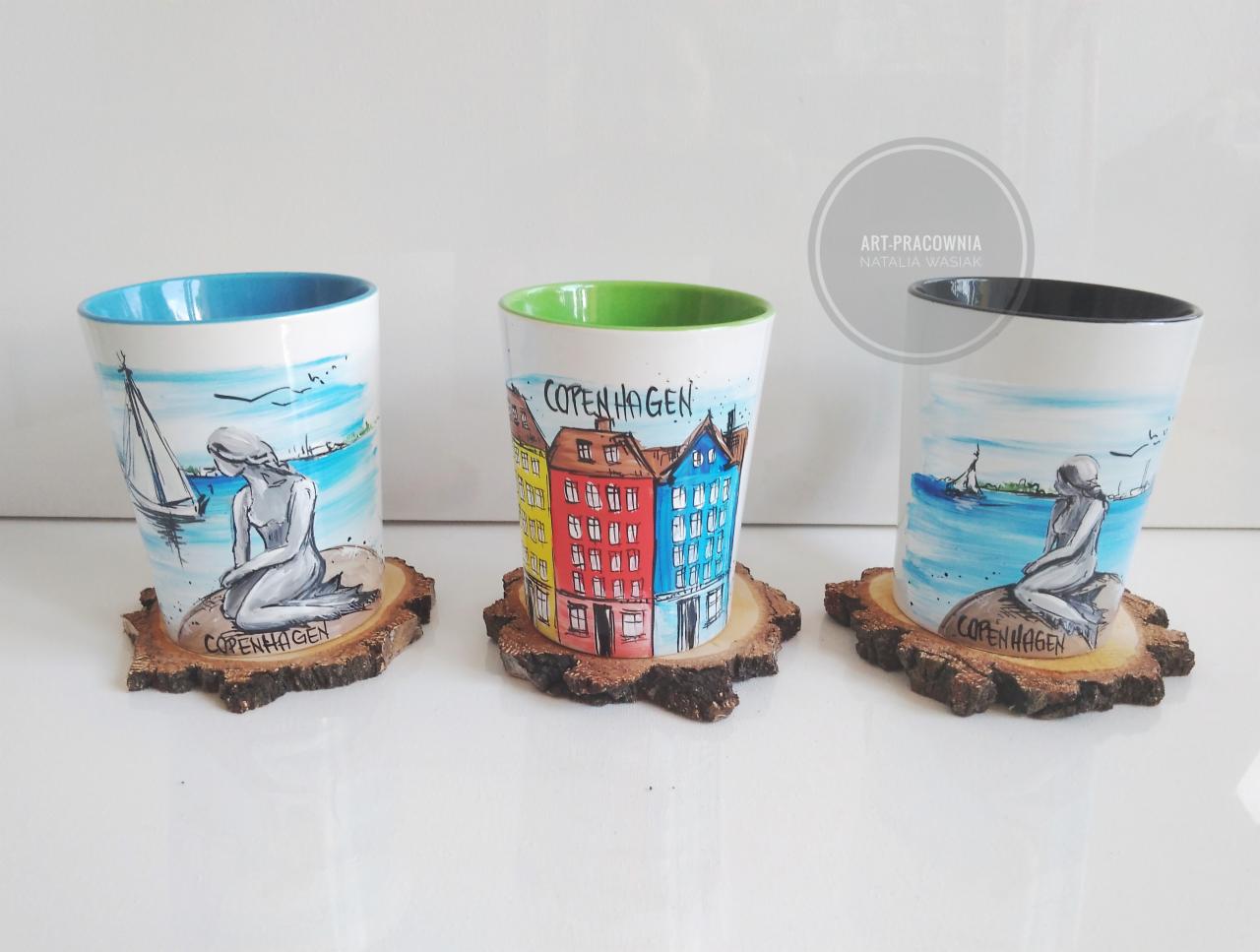 Trzy ceramiczne kubki z ręcznie malowanymi motywami Kopenhagi (syrenka, żaglówka, kolorowe kamienice), ustawione na drewnianych podkładkach imitujących plaster drewna, na białym tle.