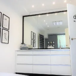 Wnętrze gabinetu z białymi meblami, lustrem w czarnej ramie i grafikami na ścianie. Na drzwiach widoczne logo: dr Kasela - medycyna estetyczna.