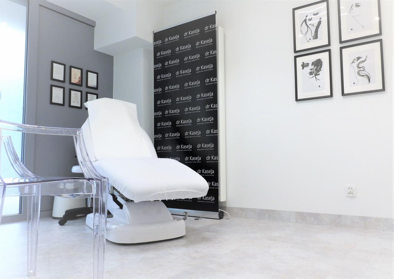 Jasne wnętrze gabinetu z fotelem zabiegowym, przezroczystymi krzesłami, czarno-białym banerem z powtarzającym się logo firmy i minimalistycznymi grafikami na ścianie.