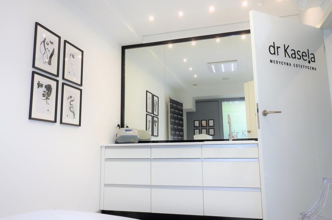 Wnętrze gabinetu z białymi meblami, lustrem w czarnej ramie i grafikami na ścianie. Na drzwiach widoczne logo: dr Kasela - medycyna estetyczna.