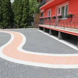 Zakręcona ścieżka z kostki brukowej w trzech kolorach: szarym, białym i łososiowym, prowadząca od furtki do domu z balkonem ozdobionym kwiatami w donicach. W tle widoczni pracownicy podczas...