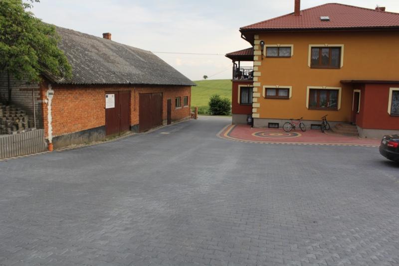 Szara kostka brukowa ułożona na podjeździe między budynkiem gospodarczym z cegły a dwukondygnacyjnym domem z żółtą elewacją, rowery zaparkowane przy ścianie.