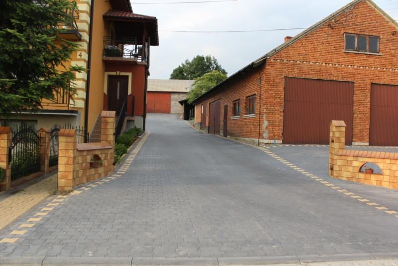 Szeroki podjazd z szarej kostki brukowej prowadzi pomiędzy budynkami, z ozdobnymi, ceglanymi murkami po bokach.