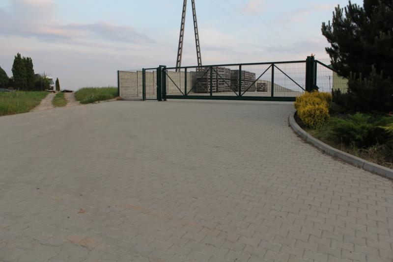 Szeroka, przesuwna brama wjazdowa z metalową kratą, za którą składowane są materiały budowlane, widoczna z perspektywy brukowanej drogi dojazdowej, z elementami zieleni po bokach.