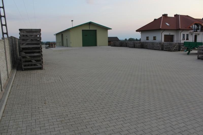 Szara kostka brukowa ułożona na dużej powierzchni placu, z budynkami w tle i stertą drewnianych skrzyń po lewej stronie.