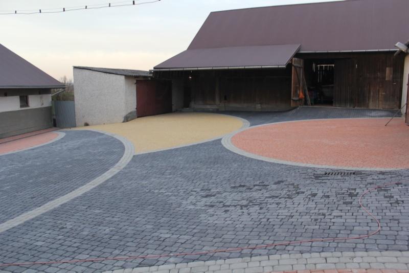 Podjazd z kostki brukowej ułożonej w okrągłe wzory z wykorzystaniem kostki w kolorze szarym, żółtym i czerwonym, na tle zabudowań gospodarczych.
