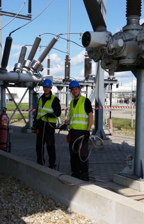 Dwóch instalatorów energetycznych w kaskach i kamizelkach odblaskowych, z przewodami pomiarowymi, stoi na terenie stacji transformatorowej pod konstrukcją wsporczą z izolatorami.