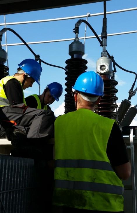 Trzech monterów w kaskach i kamizelkach odblaskowych pracuje przy transformatorze na tle błękitnego nieba.