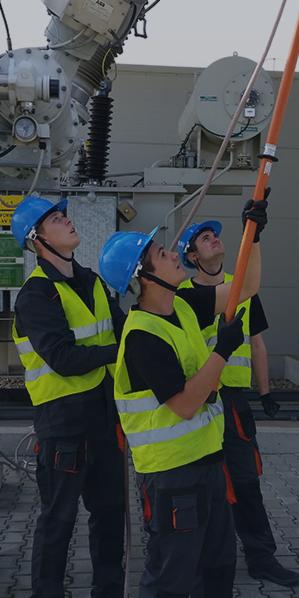 Trzech monterów w kaskach i kamizelkach odblaskowych, spoglądających w górę na transformator i używających tyczki izolacyjnej.