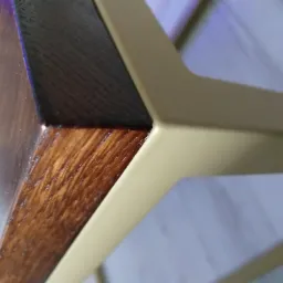 Detal narożnika blatu stołu z ciemnego drewna z widoczną strukturą słojów, połączonego z metalową nogą w kolorze jasnego złota o geometrycznym kształcie.