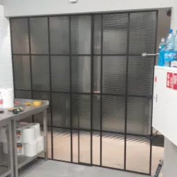 Przeszklona ścianka działowa z czarnymi metalowymi profilami, wykonana ze szkła ornamentowego, widoczna w przestrzeni przemysłowej z elementami wyposażenia.