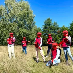 Grupa dzieci w czerwonych hełmach i kamizelkach ochronnych, trzymających czerwone pistolety do paintballa, stoi na łące w słoneczny dzień.
