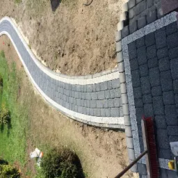 Zakrzywiony chodnik z szarej kostki brukowej z białymi obrzeżami, przechodzący przez trawnik, widoczne narzędzia brukarskie: szczotka i szpachelka.