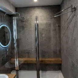 Nowoczesna łazienka z szarymi płytkami imitującymi kamień, okrągłe lustra z podświetleniem LED, kabina prysznicowa z drewnianą półką wewnątrz oraz biały grzejnik drabinkowy.