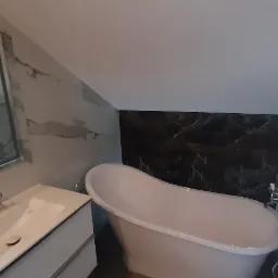 Wykończona łazienka na poddaszu z wolnostojącą wanną, umywalką z szafką i lustrem w ramie, ściany wyłożone płytkami imitującymi marmur i czarny kamień