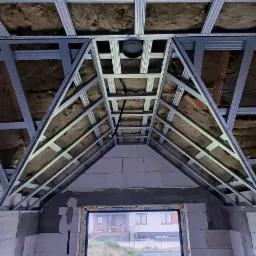 Montaż metalowego stelaża sufitu podwieszanego z izolacją z wełny mineralnej, widok od dołu na okno i niezabezpieczone ściany z pustaków.