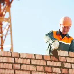 Murarz w pomarańczowym kasku układa czerwoną cegłę, w tle fragment żurawia budowlanego i błękitne niebo.