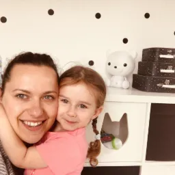 Uśmiechnięta kobieta obejmuje małą dziewczynkę z warkoczem w pokoju dziecięcym z białymi meblami i dekoracjami w kropki na ścianie.