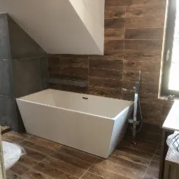 Biała, prostokątna wanna wolnostojąca w łazience na poddaszu, z brązowymi płytkami imitującymi drewno na ścianie i podłodze, oraz szarymi płytkami na ścianie po lewej stronie, widoczne elementy...